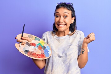 Young woman holding paintbrush and palette screaming proud, celebrating victory and success very excited with raised arm