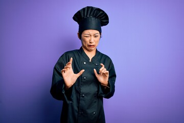Young beautiful chinese chef woman wearing cooker uniform and hat over purple background disgusted...