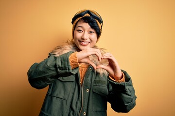 Beautiful asian skier girl wearing snow sportswear using ski goggles over yellow background smiling in love doing heart symbol shape with hands. Romantic concept.