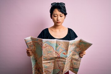 Young beautiful asian tourist girl on vacation holding city map over isolated pink background with a confident expression on smart face thinking serious © Krakenimages.com