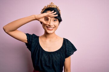 Young beautiful chinese woman wearing golden crown of king over isolated pink background doing ok gesture with hand smiling, eye looking through fingers with happy face.