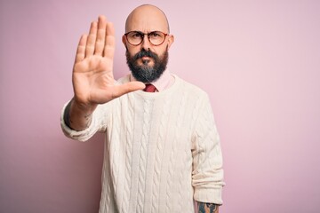 Handsome bald man with beard and tattoo wearing glasses and sweater over pink background doing stop...