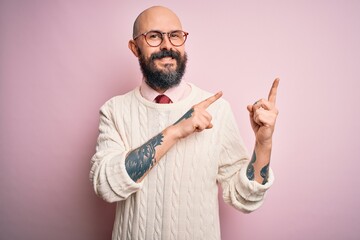 Handsome bald man with beard and tattoo wearing glasses and sweater over pink background smiling and looking at the camera pointing with two hands and fingers to the side.