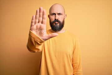 Handsome bald man with beard wearing casual sweater standing over yellow background doing stop sing...