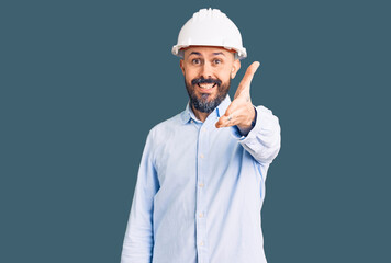 Young handsome man wearing architect hardhat smiling friendly offering handshake as greeting and welcoming. successful business.