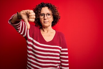 Fototapeta premium Middle age beautiful curly hair woman wearing casual striped sweater over red background looking unhappy and angry showing rejection and negative with thumbs down gesture. Bad expression.
