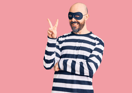 Young handsome man wearing burglar mask smiling with happy face winking at the camera doing victory sign with fingers. number two.
