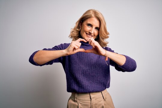 Middle age beautiful blonde woman wearing purple turtleneck sweater over white background smiling in love showing heart symbol and shape with hands. Romantic concept.