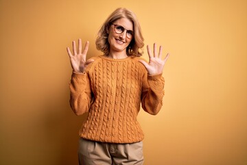 Middle age beautiful blonde woman wearing casual sweater and glasses over yellow background showing and pointing up with fingers number ten while smiling confident and happy.