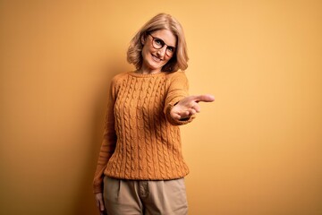 Middle age beautiful blonde woman wearing casual sweater and glasses over yellow background smiling...