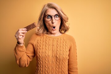 Middle age beautiful blonde woman eating energetic protein bar over yellow background scared in...