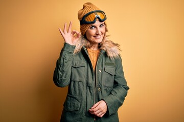 Middle age beautiful blonde skier woman wearing snow sportwear and ski goggles smiling positive doing ok sign with hand and fingers. Successful expression.