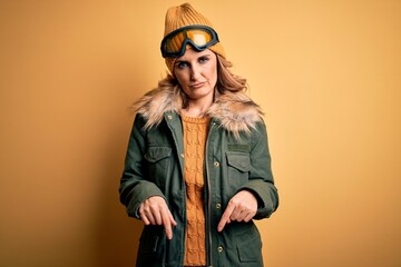 Middle age beautiful blonde skier woman wearing snow sportwear and ski goggles Pointing down looking sad and upset, indicating direction with fingers, unhappy and depressed.