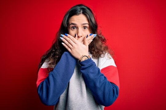 Young beautiful woman with curly hair wearing casual sweatshirt over isolated red background shocked covering mouth with hands for mistake. Secret concept.