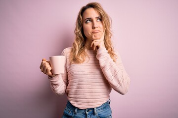 Young beautiful blonde woman drinking mug of coffee over isolated pink background serious face...