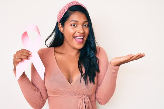 Beautiful latin young woman with long hair holding pink cancer ribbon celebrating achievement with happy smile and winner expression with raised hand