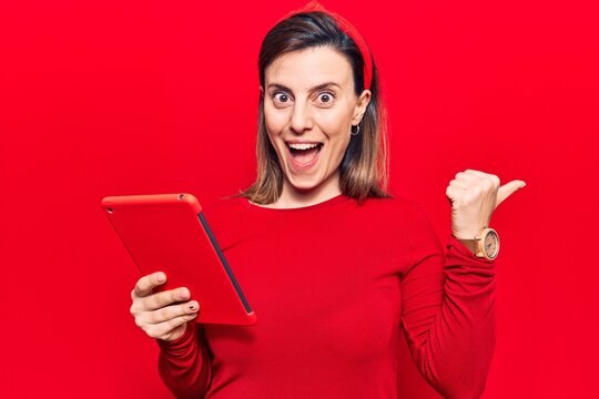 Young beautiful woman holding touchpad pointing thumb up to the side smiling happy with open mouth