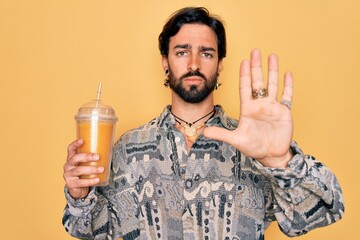 Young handsome hispanic bohemian hipster man with bear drinking healthy fresh orange juice with...