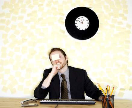 A man in business suit sitting at his desk with a stick-on paper on his forehead