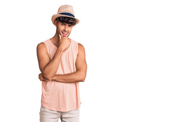 Young hispanic man wearing summer hat looking confident at the camera smiling with crossed arms and hand raised on chin. thinking positive.