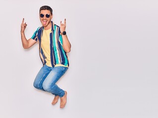 Young handsome hispanic man wearing casual clothes and sunglasses smiling happy. Jumping with smile on face doing horns sign with fingers over isolated white background