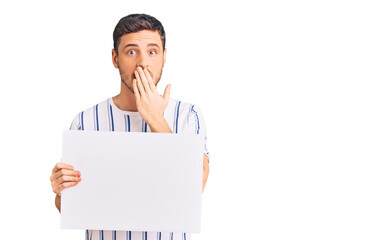Handsome young man with bear holding blank empty banner covering mouth with hand, shocked and...