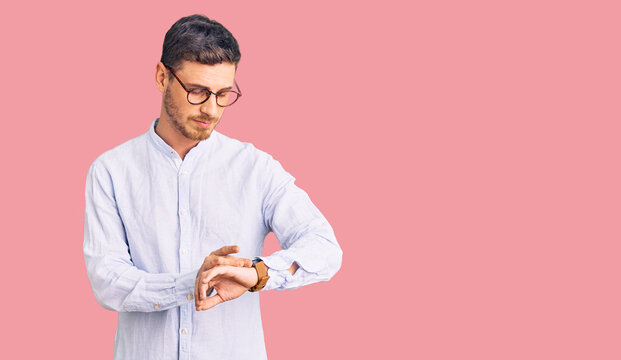 Handsome young man with bear wearing elegant business shirt and glasses checking the time on wrist watch, relaxed and confident