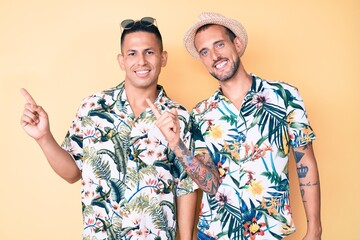Young gay couple of two men wearing summer hat and hawaiian shirt smiling and looking at the camera pointing with two hands and fingers to the side.