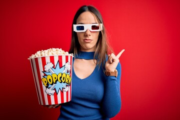 Obraz premium Young blonde woman wearing 3d glasses and eating pack of popcorn watching a movie on cinema Pointing with hand finger to the side showing advertisement, serious and calm face