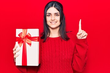 Young beautiful girl holding gift smiling with an idea or question pointing finger with happy face, number one
