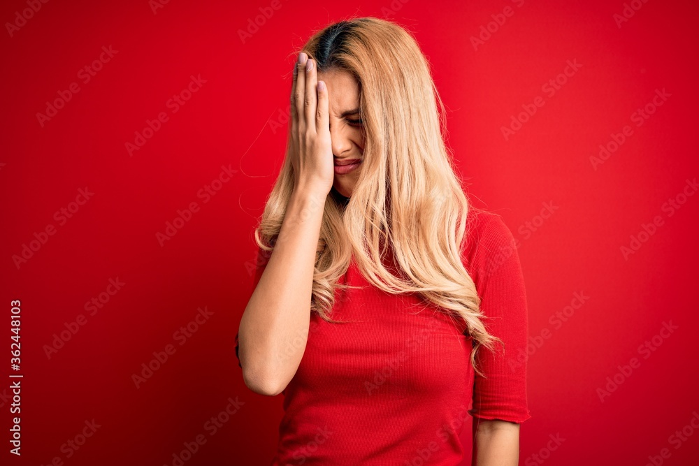 Wall mural Young beautiful blonde woman wearing casual t-shirt standing over isolated red background Yawning tired covering half face, eye and mouth with hand. Face hurts in pain.