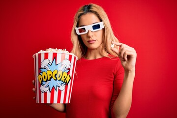 Obraz premium Young beautiful blonde woman watching movie using 3d glasses eating popcorns as snack with a confident expression on smart face thinking serious