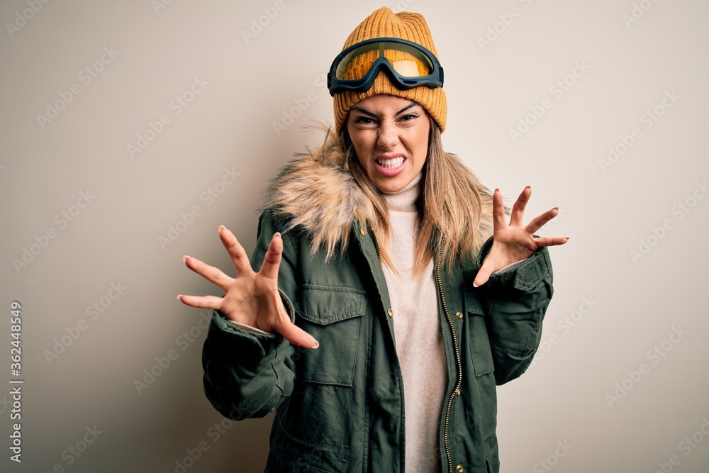 Poster Young brunette skier woman wearing snow clothes and ski goggles over white background smiling funny doing claw gesture as cat, aggressive and sexy expression