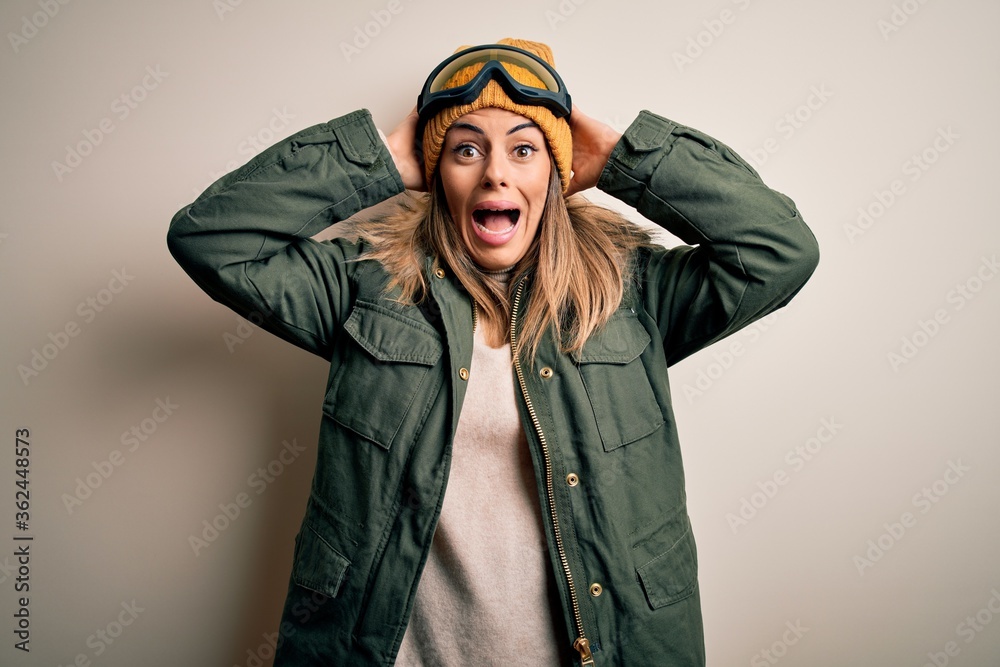 Poster Young brunette skier woman wearing snow clothes and ski goggles over white background Crazy and scared with hands on head, afraid and surprised of shock with open mouth