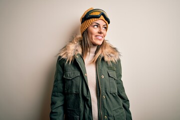 Young brunette skier woman wearing snow clothes and ski goggles over white background looking away to side with smile on face, natural expression. Laughing confident.