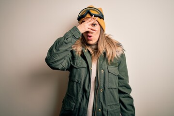 Young brunette skier woman wearing snow clothes and ski goggles over white background peeking in shock covering face and eyes with hand, looking through fingers with embarrassed expression.