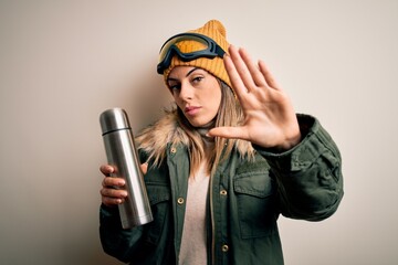 Young beautiful brunette skier woman wearing ski goggles drinking thermo with coffe with open hand doing stop sign with serious and confident expression, defense gesture