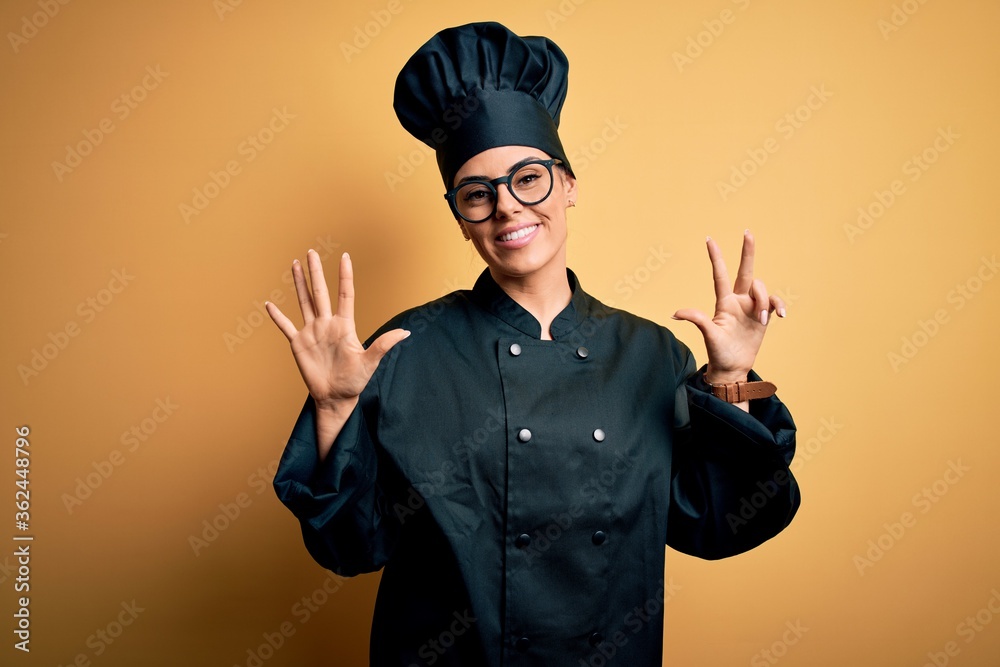 Poster Young beautiful brunette chef woman wearing cooker uniform and hat over yellow background showing and pointing up with fingers number eight while smiling confident and happy.