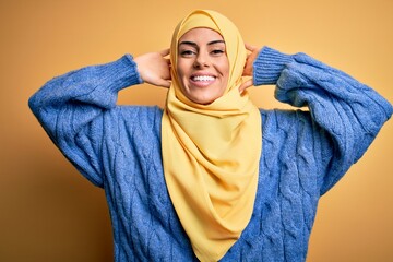 Young beautiful brunette muslim woman wearing arab hijab over isolated yellow background relaxing and stretching, arms and hands behind head and neck smiling happy