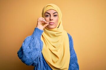 Young beautiful brunette muslim woman wearing arab hijab over isolated yellow background mouth and lips shut as zip with fingers. Secret and silent, taboo talking