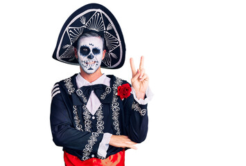 Young man wearing day of the dead costume over background smiling with happy face winking at the camera doing victory sign. number two.
