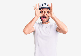 Young handsome man wearing bike helmet doing ok gesture like binoculars sticking tongue out, eyes looking through fingers. crazy expression.