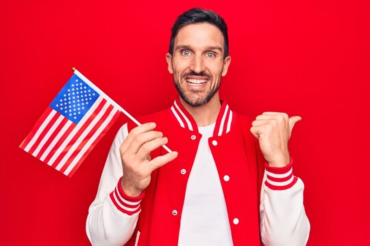 Young handsome patriotic man holding united states flag celebrating independence day pointing thumb up to the side smiling happy with open mouth
