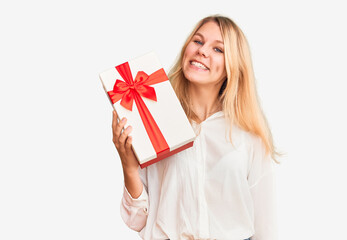 Young beautiful blonde woman holding gift looking positive and happy standing and smiling with a confident smile showing teeth