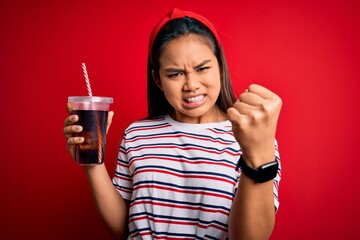 Young asian girl drinking cola fizzy refreshment using straw over isolated red background annoyed and frustrated shouting with anger, crazy and yelling with raised hand, anger concept