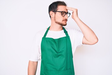 Young handsome man wearing apron surprised with hand on head for mistake, remember error. forgot,...