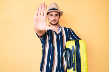 Young handsome man wearing summer hat holding cabin bag with open hand doing stop sign with serious and confident expression, defense gesture