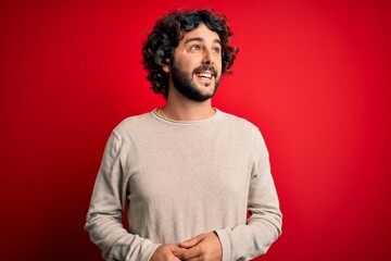Fototapeta na wymiar Young handsome man with beard wearing casual sweater standing over red background looking away to side with smile on face, natural expression. Laughing confident.