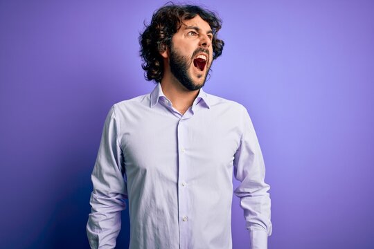 Young handsome business man with beard wearing shirt standing over purple background angry and mad screaming frustrated and furious, shouting with anger. Rage and aggressive concept.