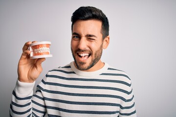 Young handsome man with beard holding plastic denture teeth over white background winking looking...